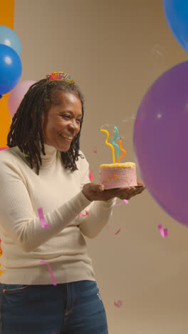 Foto-De-Estudio-De-Vídeo-Vertical-De-Una-Mujer-Con-Diadema-De-Cumpleaños-Celebrando-Un-Cumpleaños-Soplando-Velas-En-Un-Pastel-Con-Confeti-De-Papel